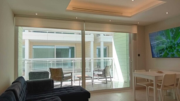 A modern living room with a dark sofa, a dining table with chairs, and a large sliding glass door leading to a balcony with outdoor furniture. - Marina Club Adriatic, Gibraltar - Studio Apartment (Gibraltar)