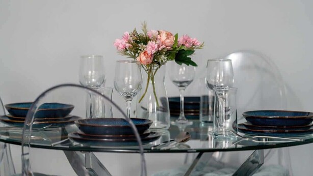 A round glass table with blue plates and clear glasses, a vase with pink flowers, and a clear glass chair. - Two-level Apartment with Terrace | Workspace (Poznan)
