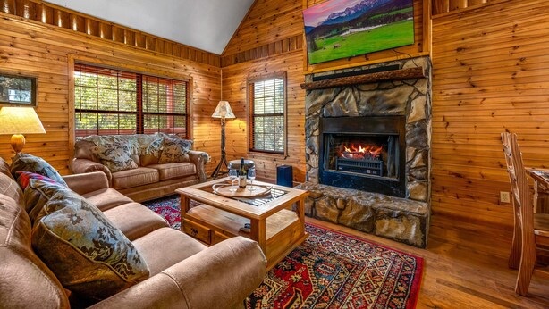 A cozy log cabin interior with a stone fireplace, leather sofas, and a large TV mounted on the wall. - Hot Tub ★ Pool Table ★ Firepit ★Very Private (Townsend)