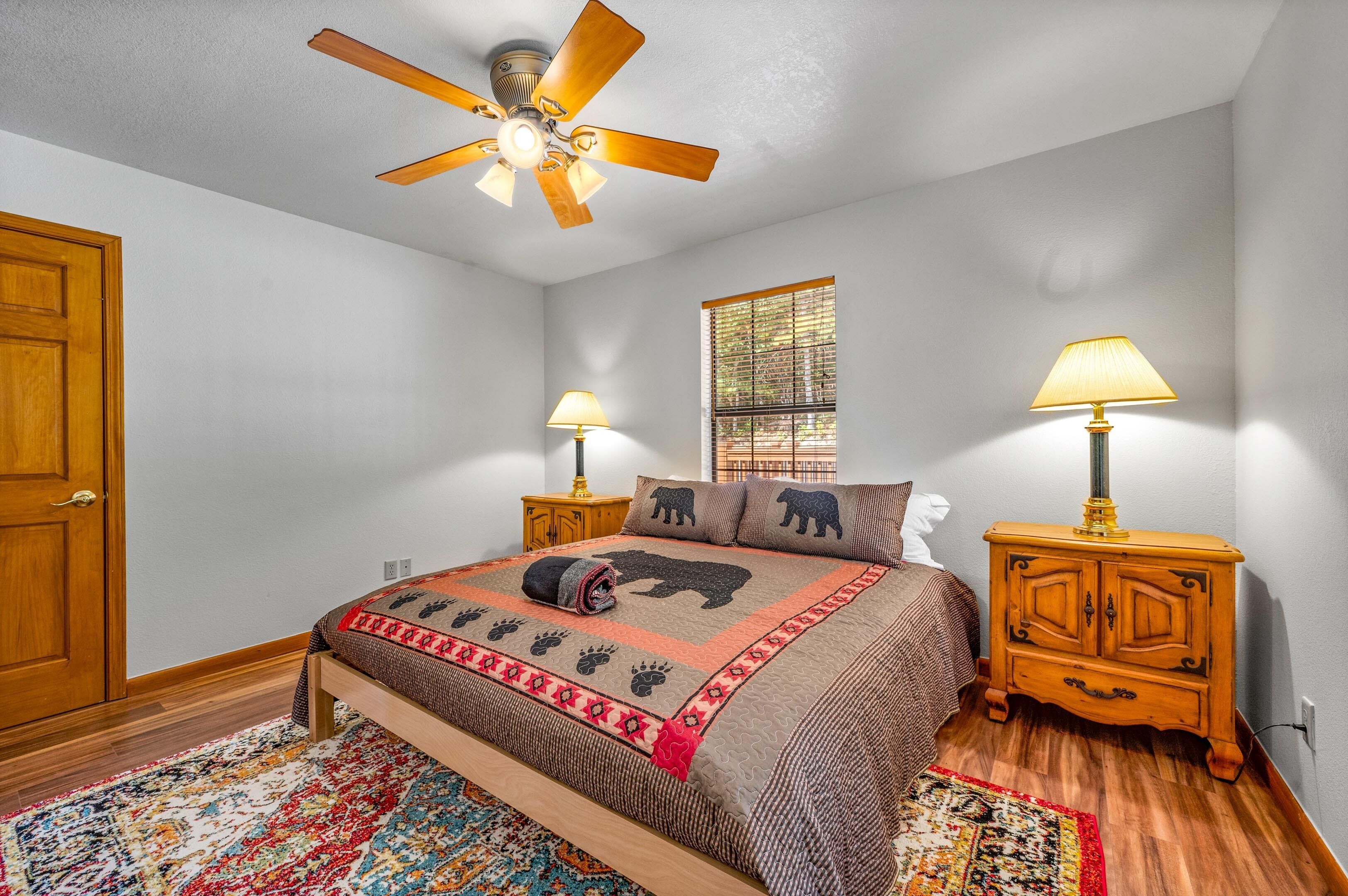 A bedroom with a bed, wooden nightstands, a ceiling fan, a window, and a door.