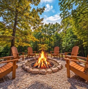 A circular fire pit with logs, surrounded by four wooden Adirondack chairs, set on a gravel surface with trees in the background.
