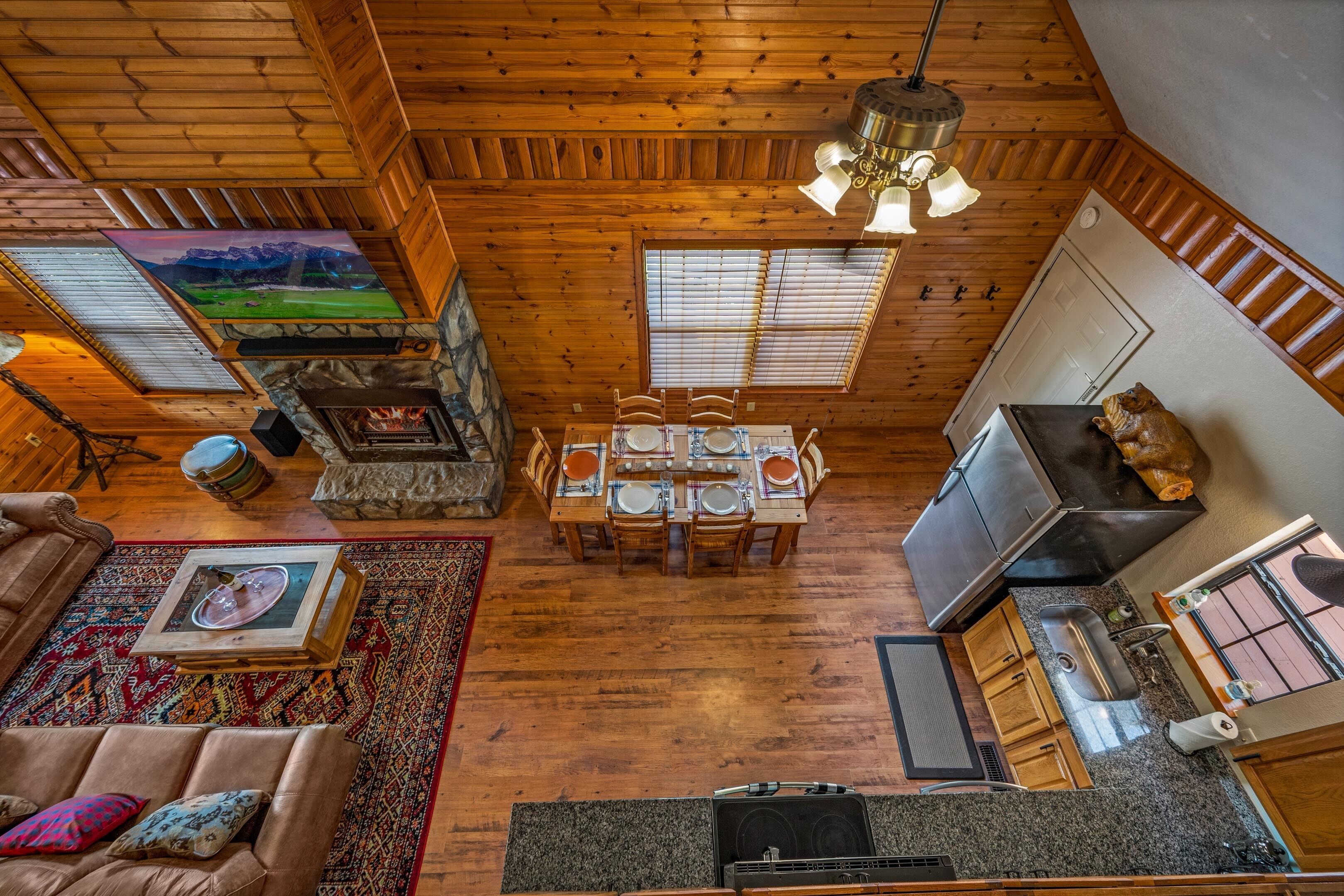 A spacious room with a wooden ceiling, a dining area with a table set for four, a fireplace, and a kitchen area with a sink and stove.