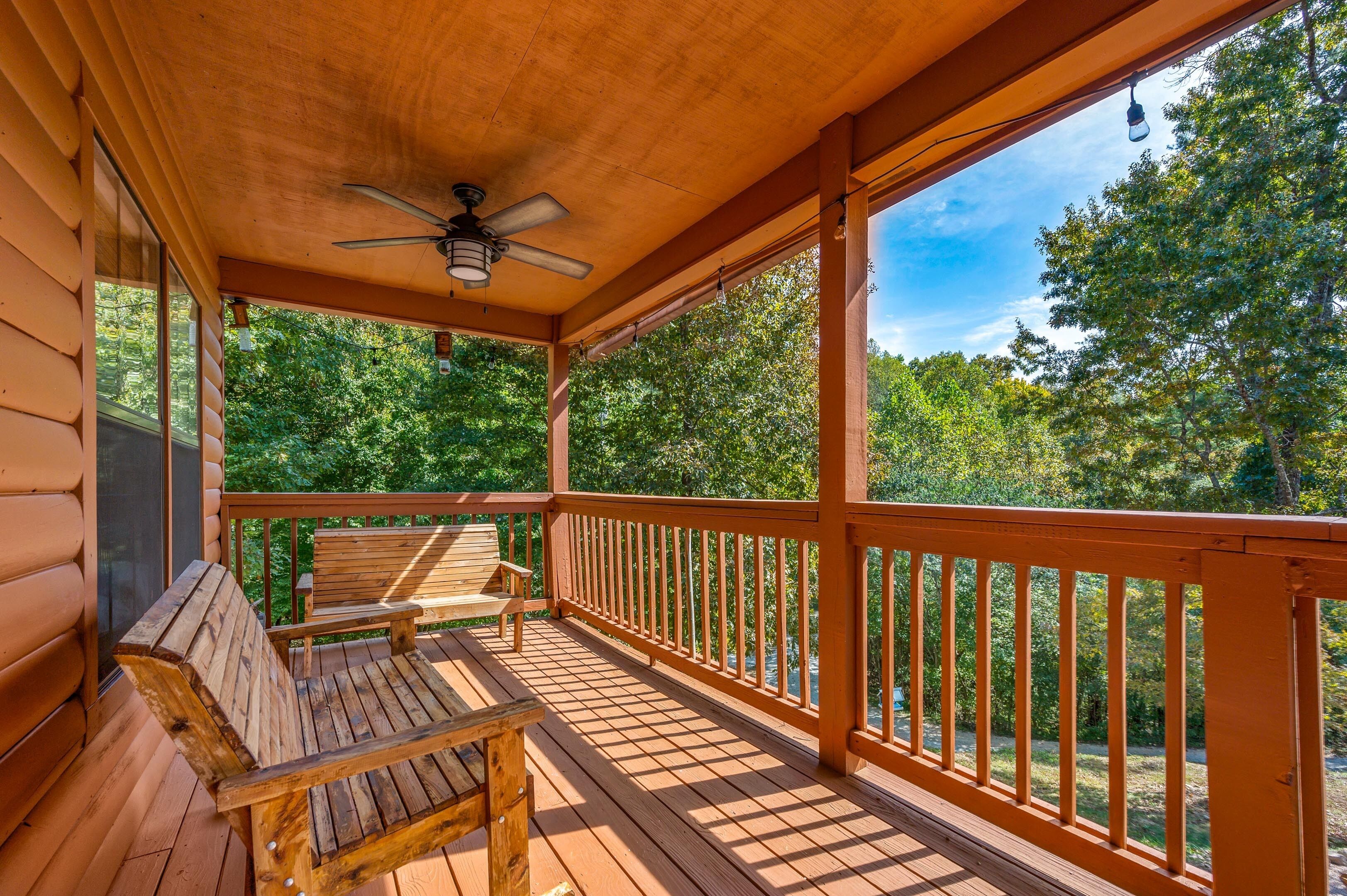 A wooden deck with a ceiling fan, wooden benches, and a railing overlooking a forested area.