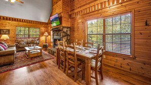 A cozy cabin interior with a wooden dining table set for four, a fireplace, and large windows offering a view of the outdoors. - Hot Tub ★ Pool Table ★ Firepit ★Very Private (Townsend)