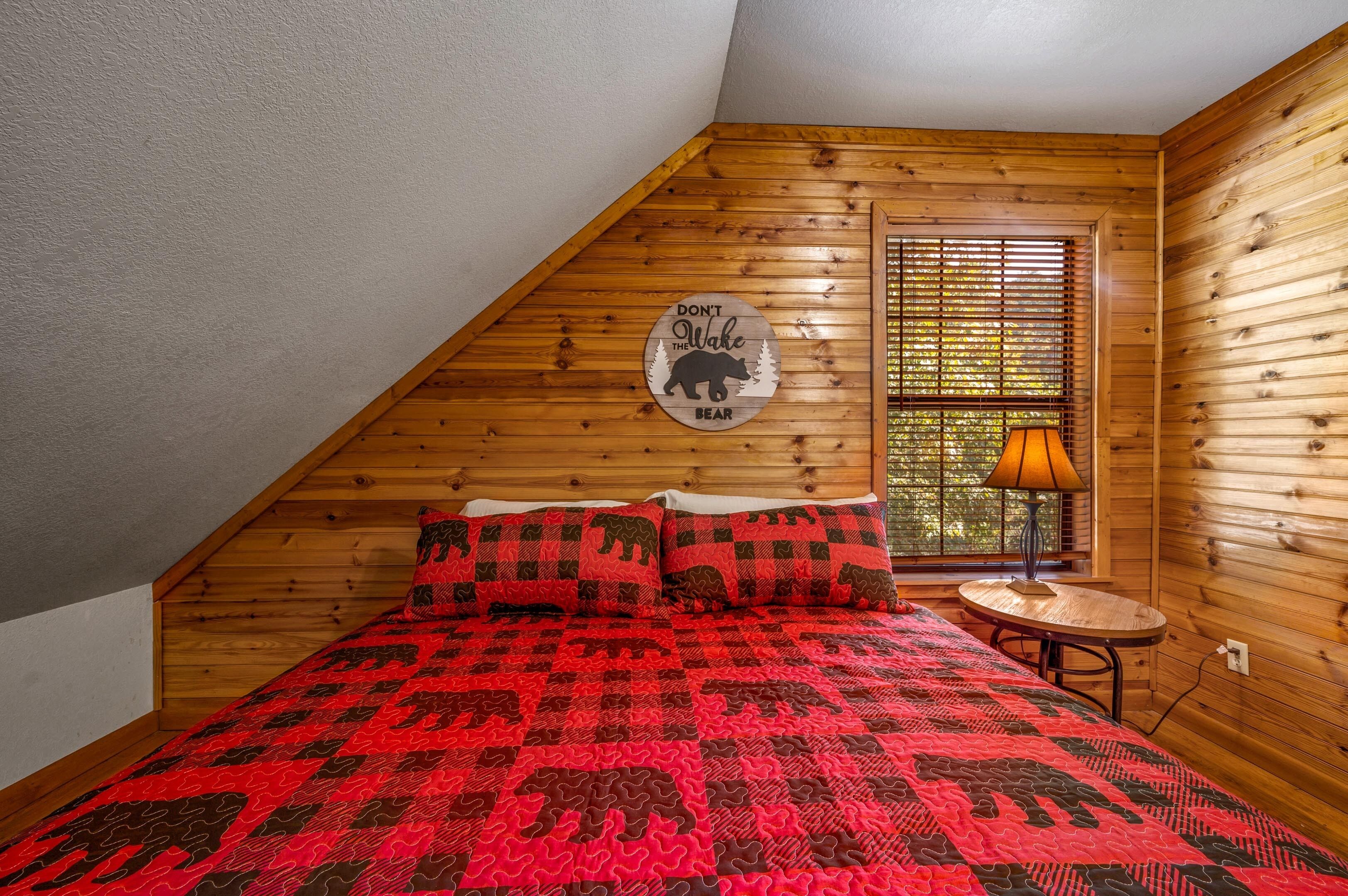 A bedroom with a sloped ceiling, a bed with a red and black patterned bedspread, two matching pillows, a small round table with a lamp, and a window with blinds.