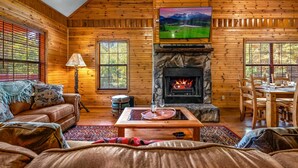 A cozy log cabin interior with a fireplace, wooden walls, a sofa, a dining table, and a television mounted above the fireplace. - Hot Tub ★ Pool Table ★ Firepit ★Very Private (Townsend)