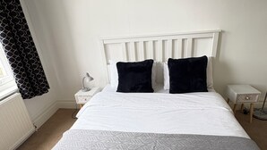 Ein Bett mit einem weißen Kopfteil, zwei schwarzen Kissen, einer grauen Decke und einem weißen Nachttisch mit einer Lampe.