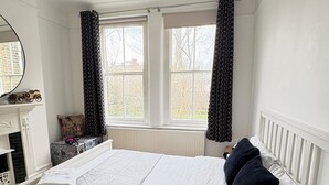 Ein Schlafzimmer mit Bett, Schminktisch, Spiegel, Fenster mit Vorhängen und Heizkörper.