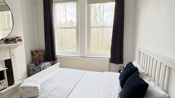 Ein Schlafzimmer mit Bett, Schminktisch, Spiegel, Fenster mit Vorhängen und Heizkörper.
