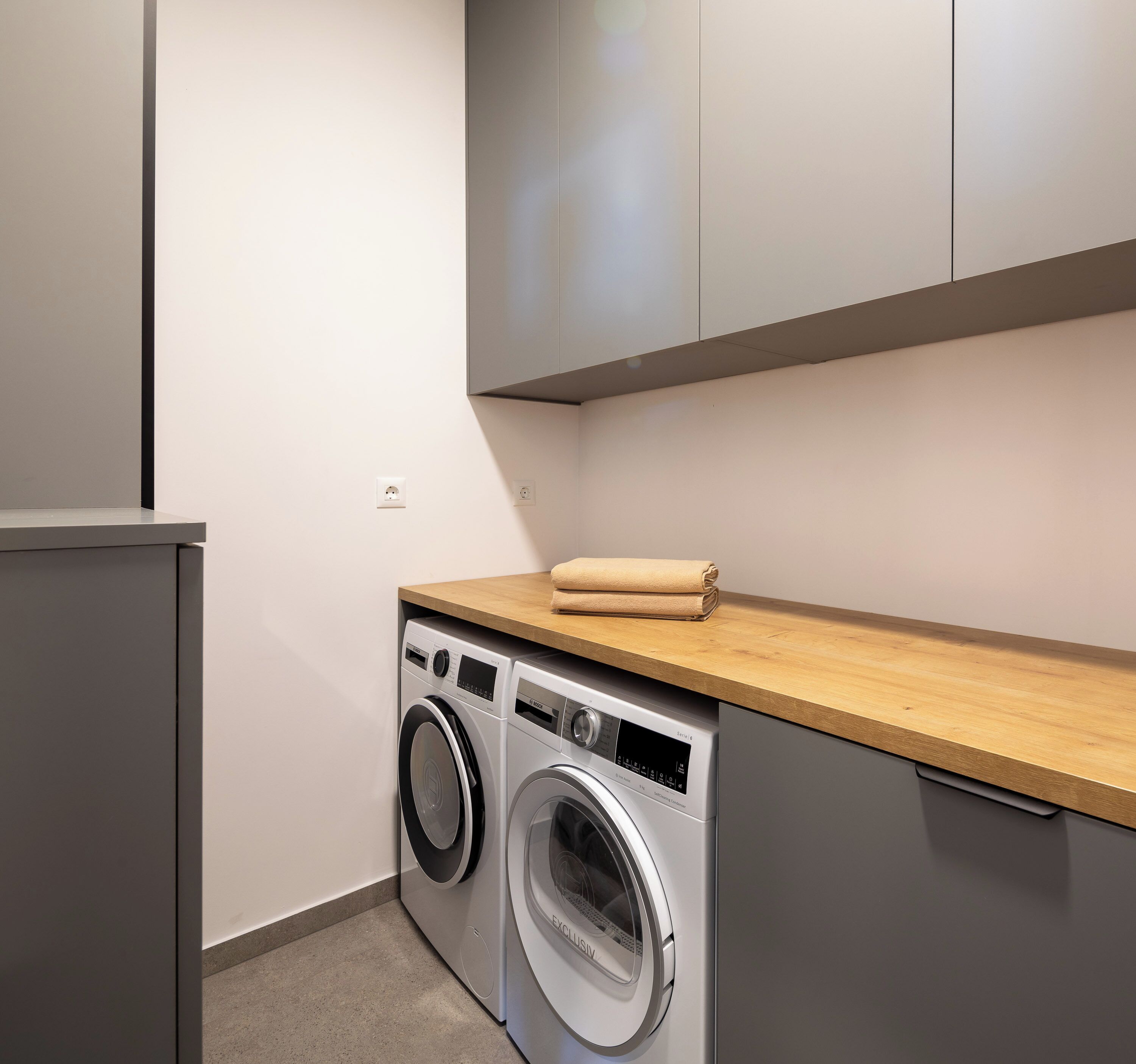 A compact laundry area with two front-loading washing machines, a wooden countertop, and a small cabinet above.