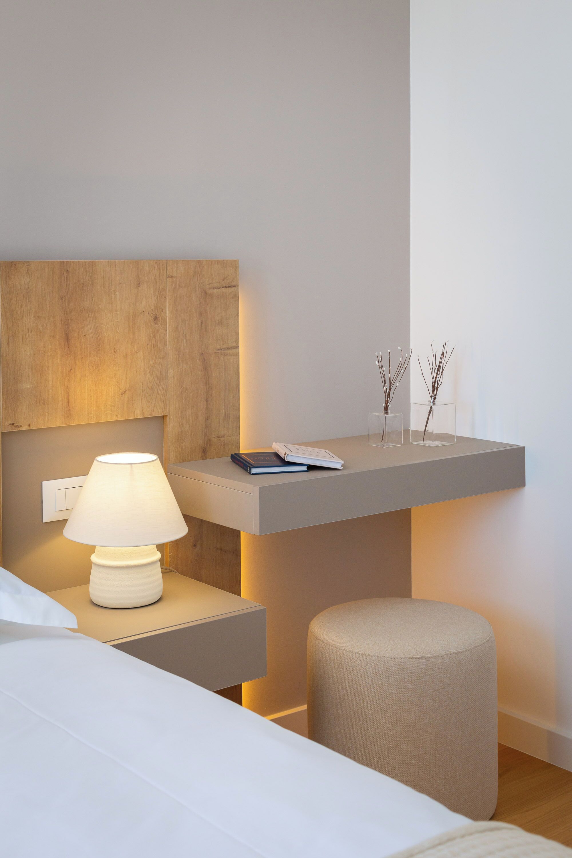 A modern bedroom with a bedside table, a lamp, a small shelf with decorative items, and a cushioned stool.