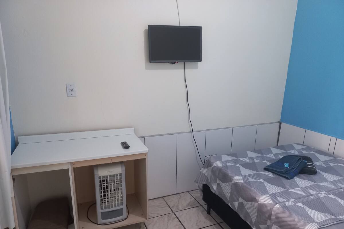 A bedroom with a bed, a TV mounted on the wall, a heater, and a small table with a remote control.