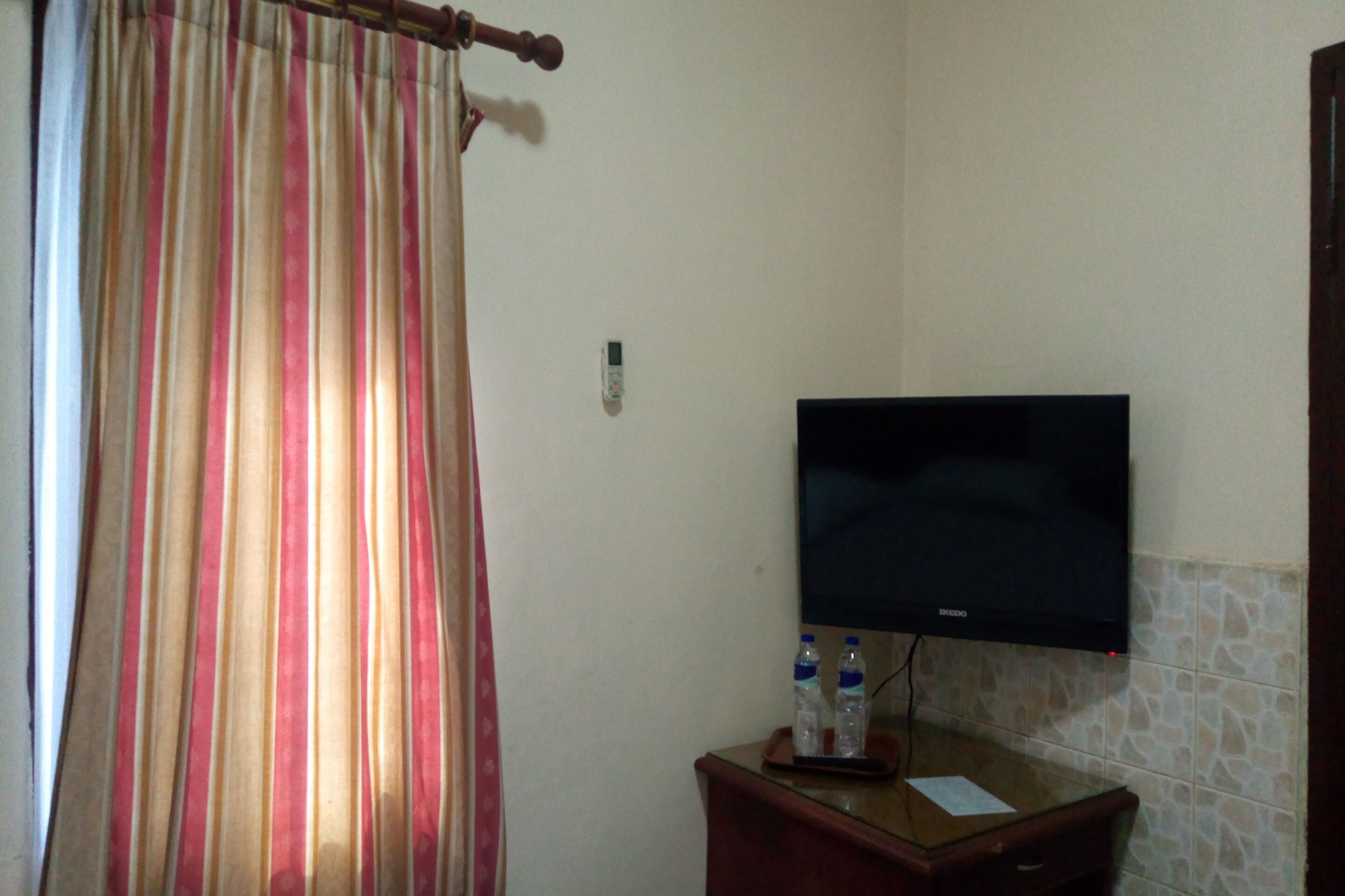 Een badkamer met een gestreept gordijn, een flatscreen televisie aan de muur en een klein houten kastje met twee waterflesjes.