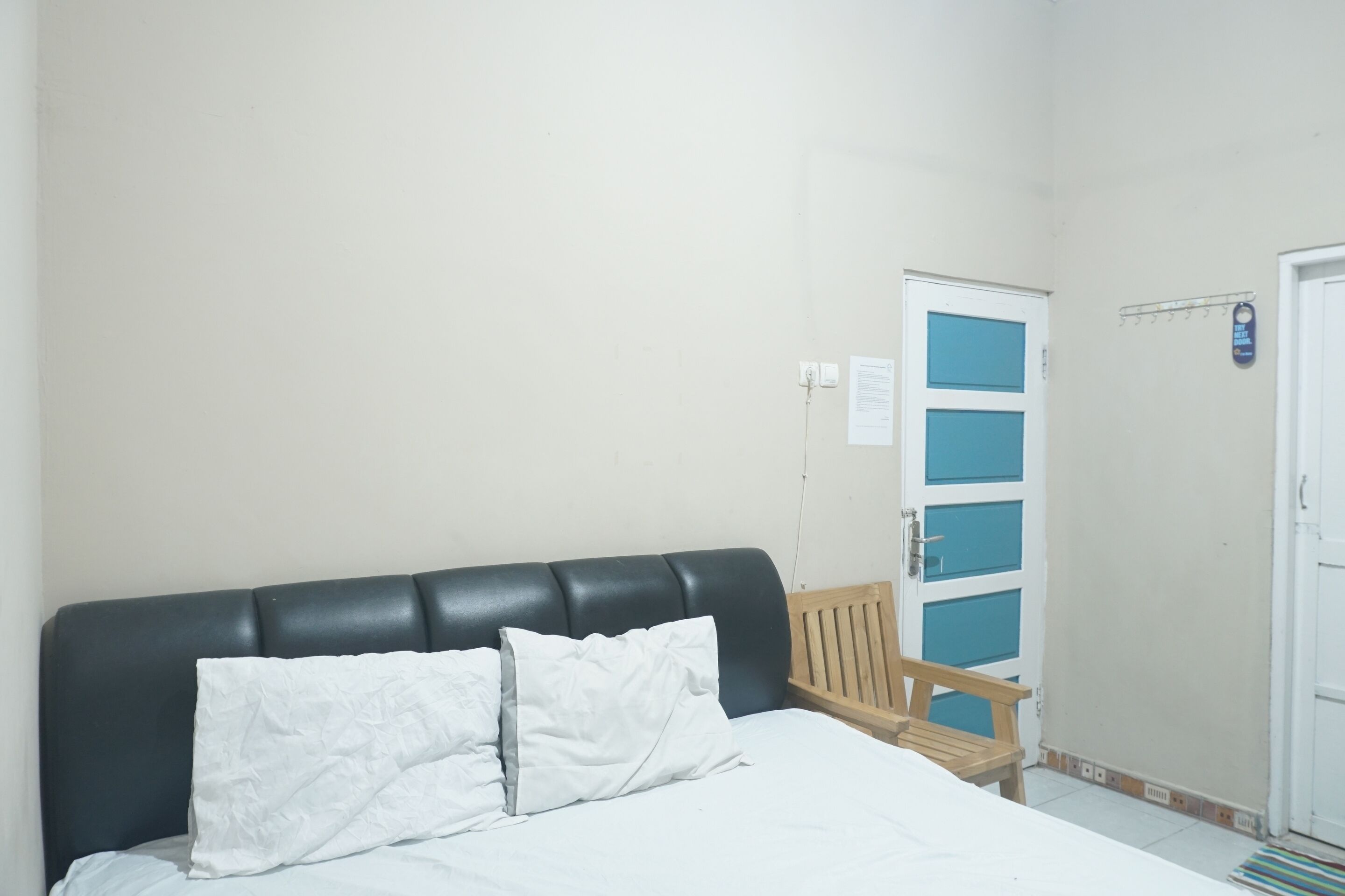 Un dormitorio con una cama, un banco de madera, una puerta blanca con paneles azules e interruptor de luz fijado a la pared.