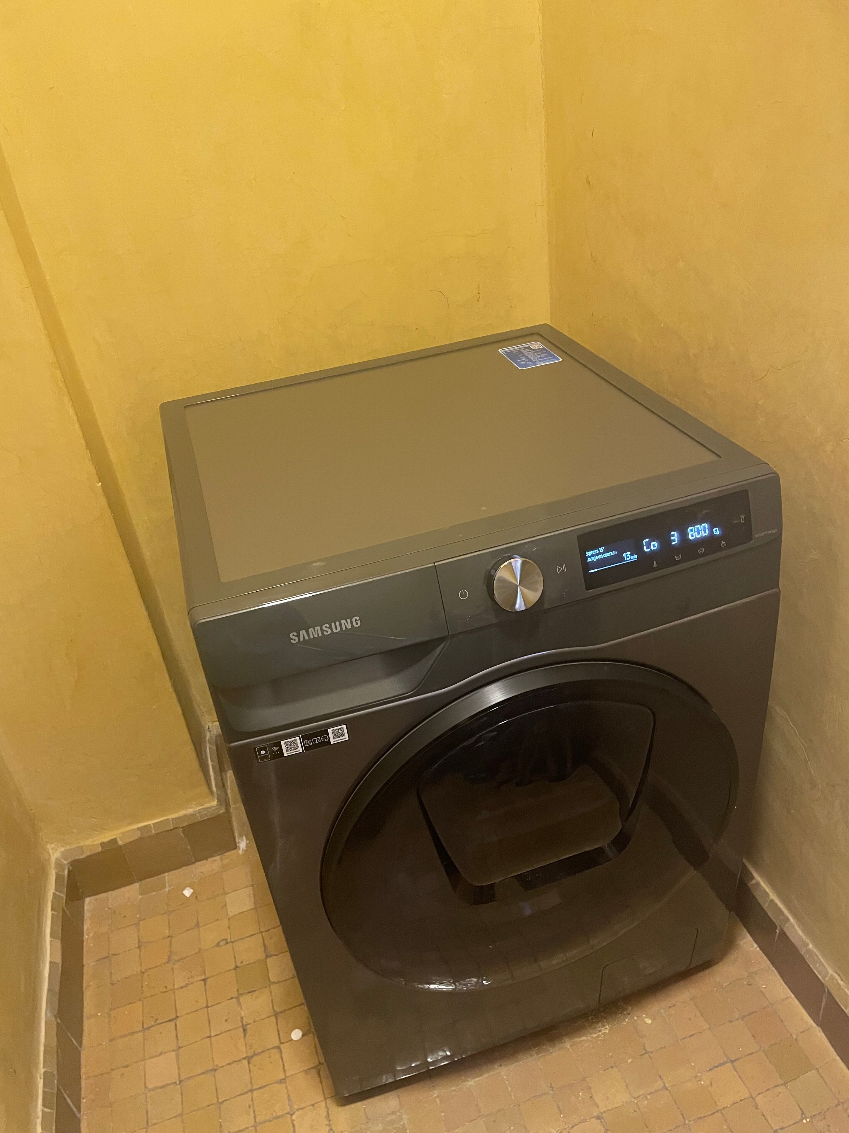Een Samsung wasmachine in een hoek van een kamer met gele wanden en een betegelde vloer.