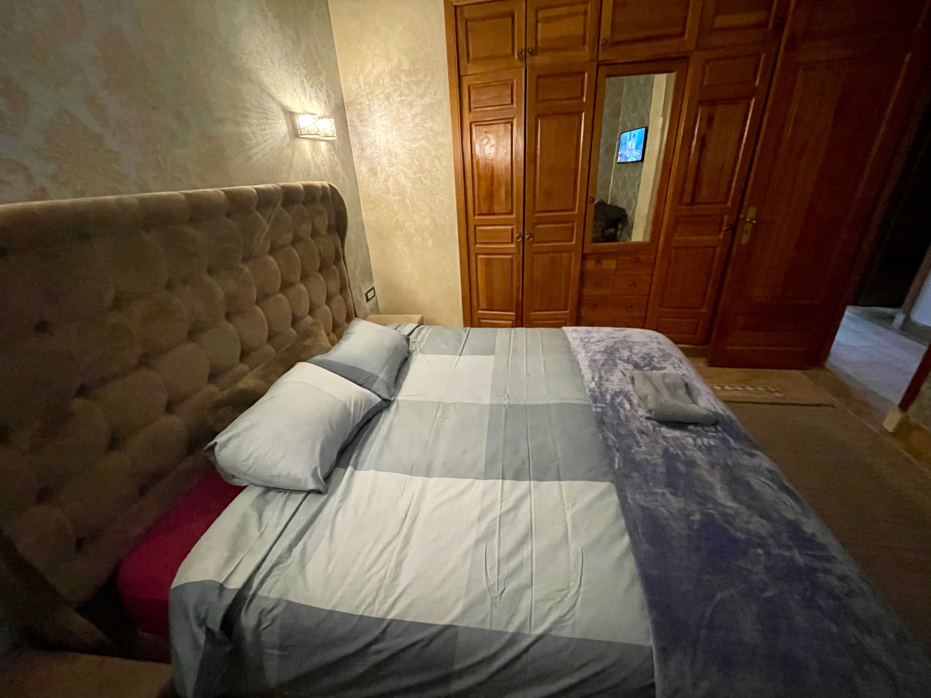 A bedroom with a tufted headboard, a bed with a grey and white striped bedspread, and wooden wardrobes.