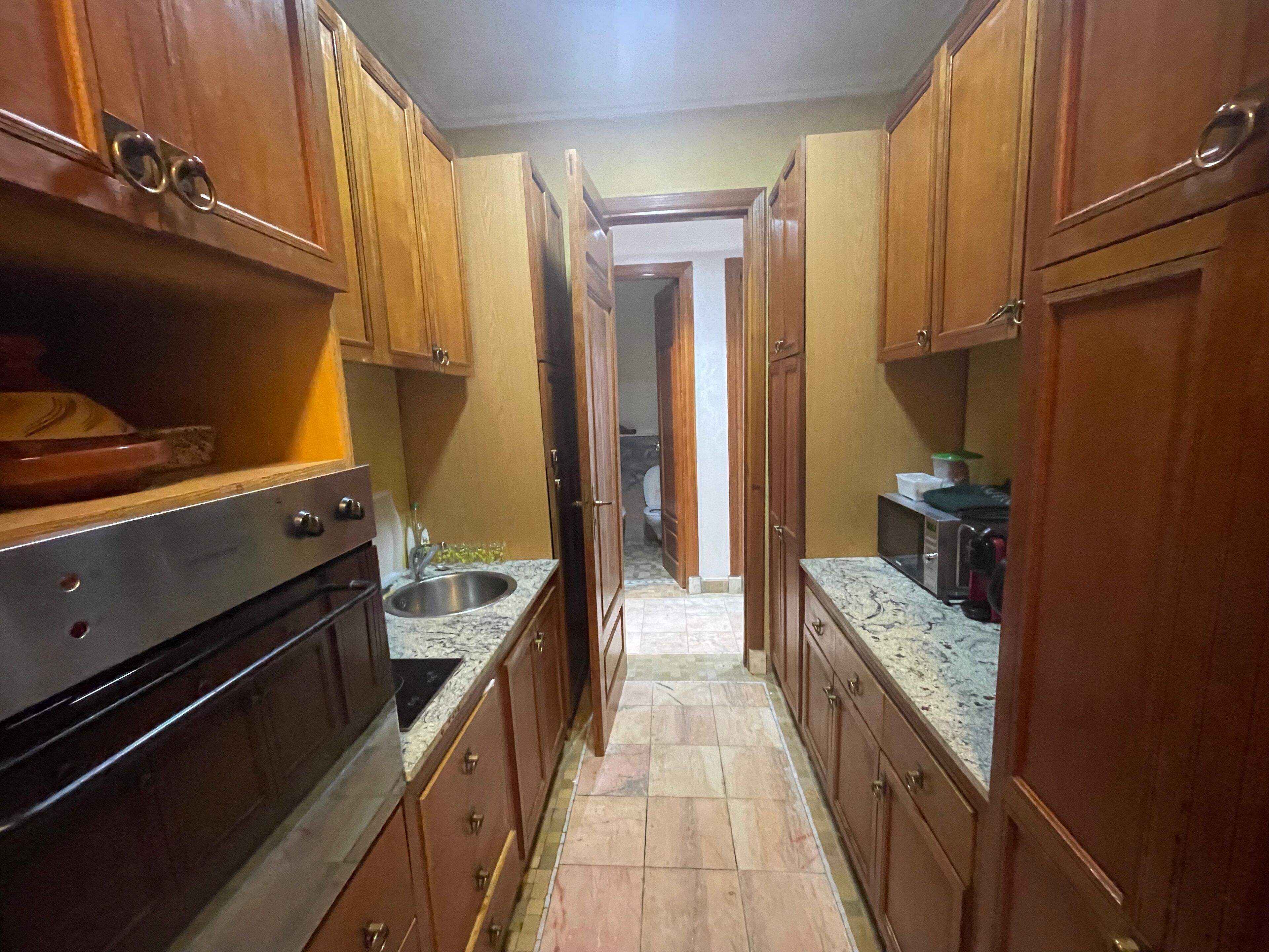 Una cocina estrecha con armarios de madera, un lavamanos de acero inoxidable y suelo de baldosas.
