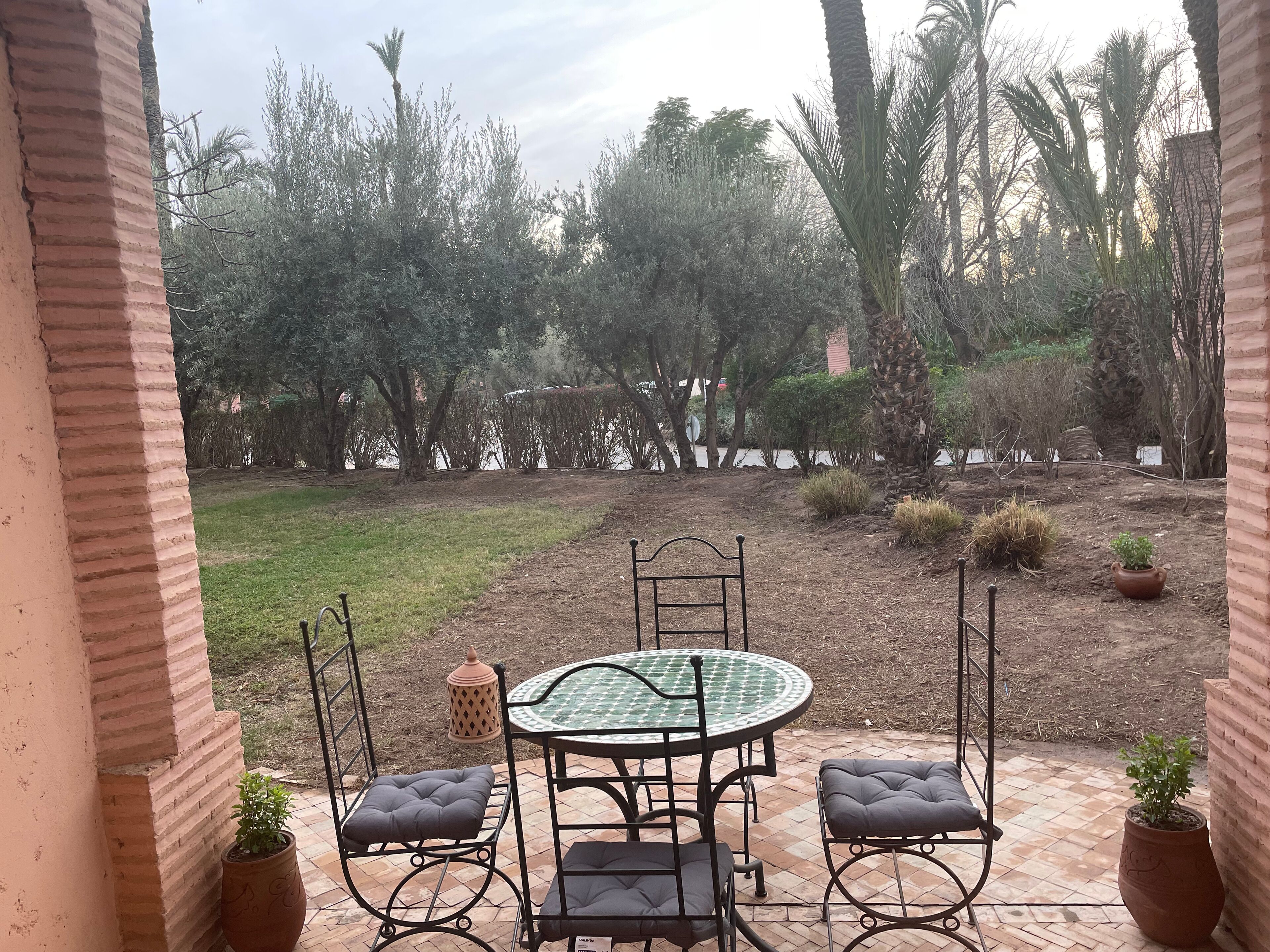 A patio with a round table and four chairs, surrounded by potted plants and a view of a landscaped garden.