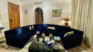 Una sala de estar con un sofá azul oscuro, una mesa de centro de vidrio y una araña.