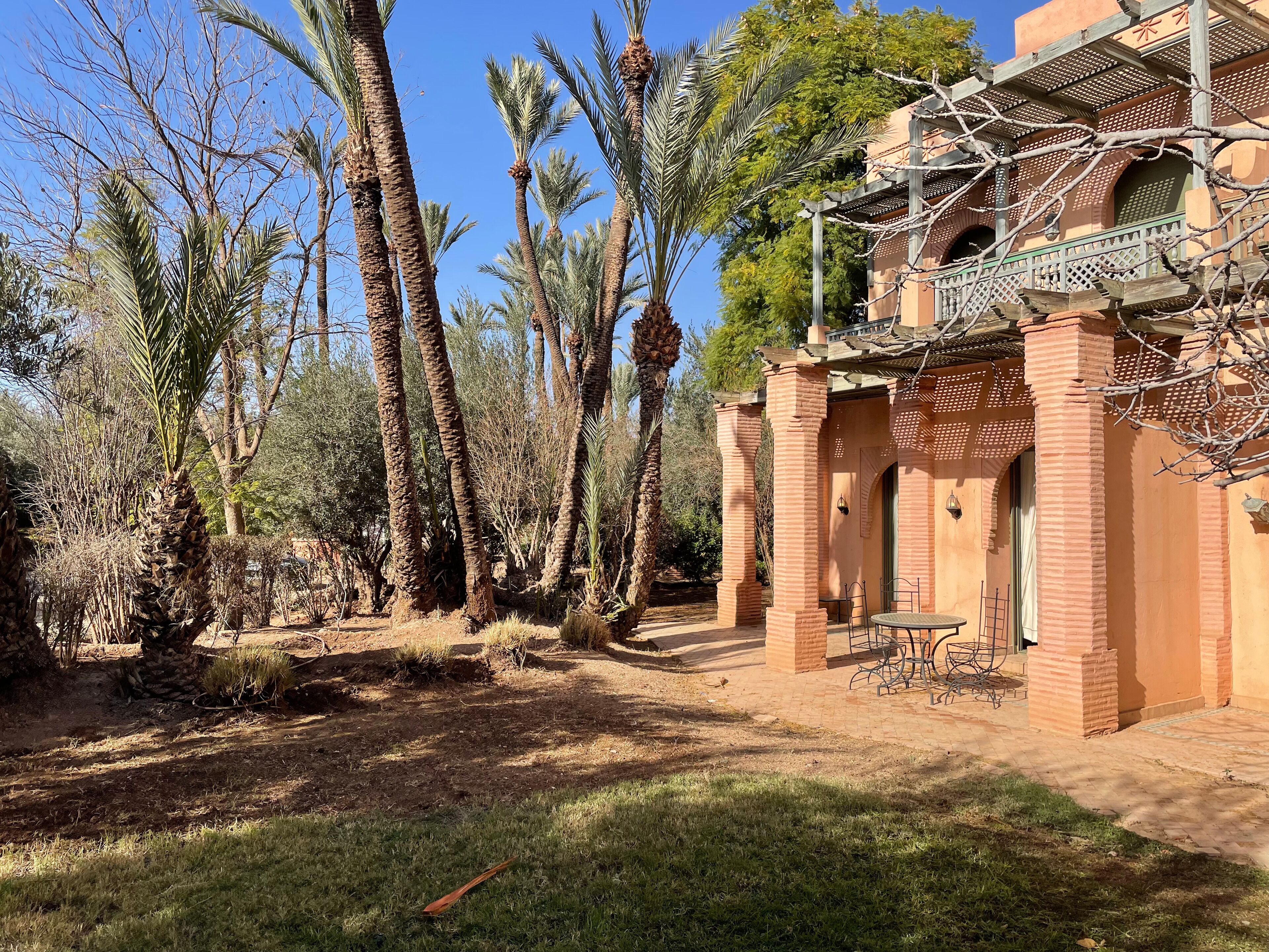 A courtyard with palm trees, a brick building with a balcony, and a small table with chairs.