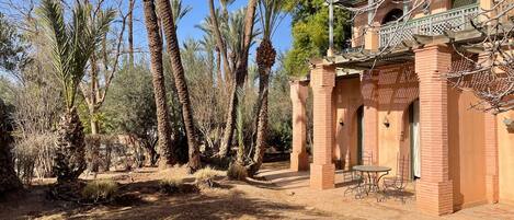 A courtyard with palm trees, a brick building with a balcony, and a small table with chairs.