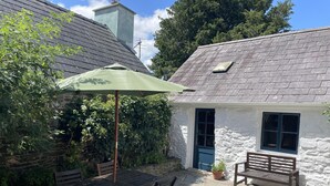 Une petite maison en pierre avec un toit de tuiles, un parasol vert, une table et des chaises en bois, et un banc.