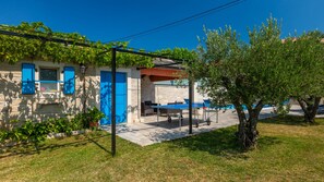 Ein Steingebäude mit blauen Fensterläden, einer mit Weinreben bewachsenen Pergola, Außenbestuhlung und einem Baum.