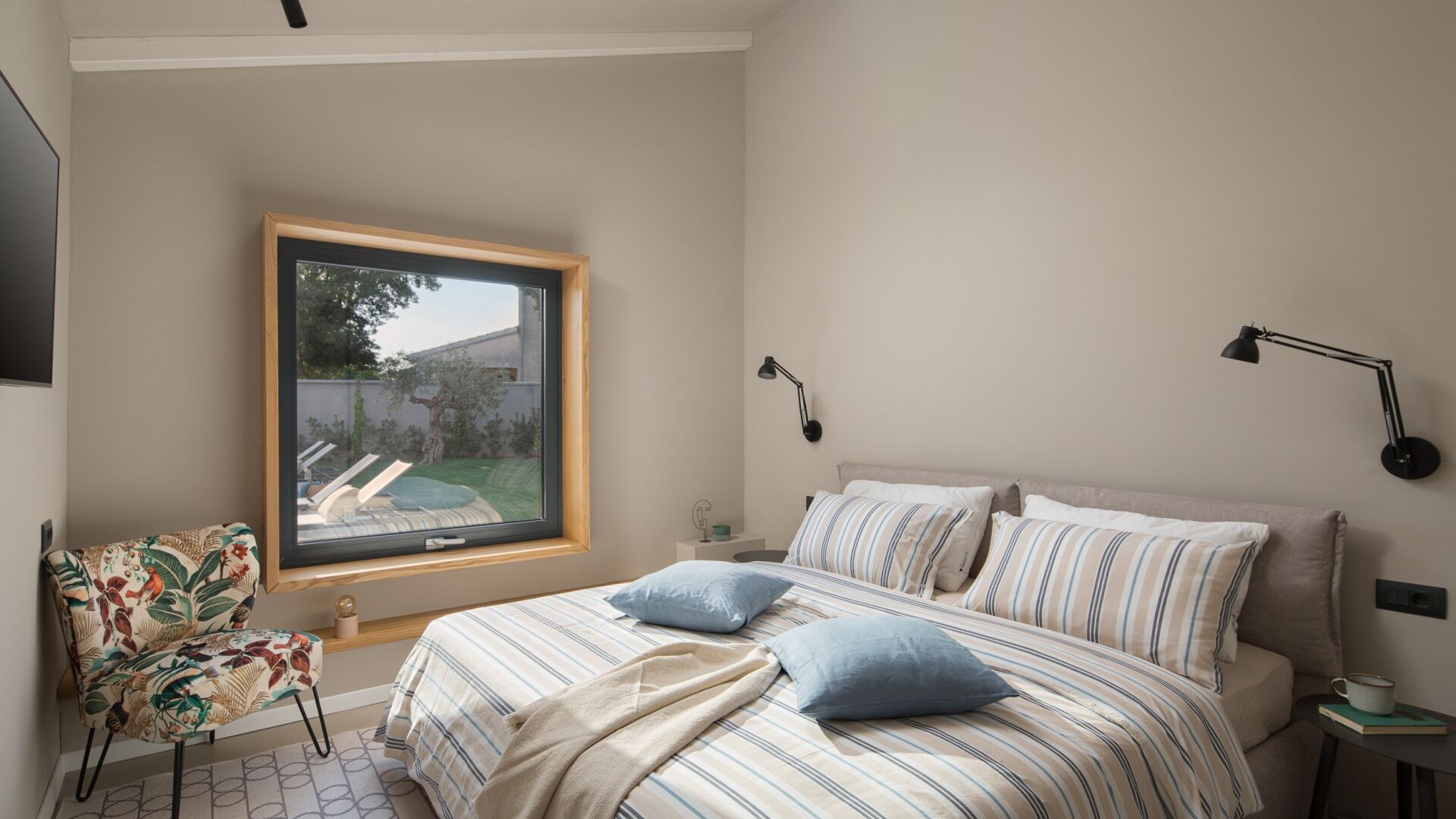 Ein Schlafzimmer mit großem Fenster, einem Bett mit gestreifter Bettwäsche, einem Sessel mit Blumenmuster und einem Nachttisch mit Lampe.