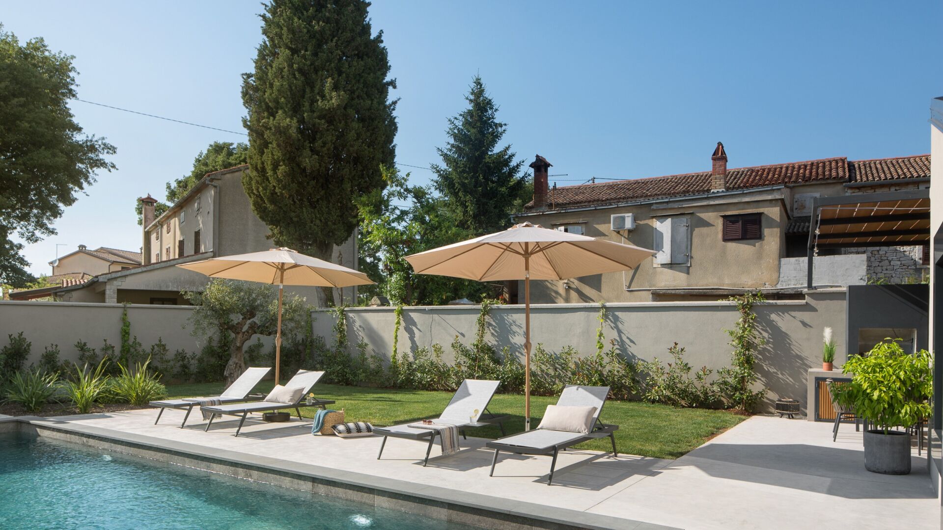 A poolside area with lounge chairs and umbrellas, surrounded by a garden and a building with a tiled roof.