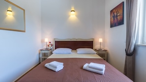 A neatly made bed with two pillows and two folded towels, two bedside lamps, a nightstand with a decorative item, and a painting on the wall.