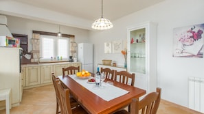 Un coin repas avec une table en bois, des chaises et un lustre. On aperçoit au fond une cuisine équipée d’un réfrigérateur, d’une cuisinière et d’un évier.