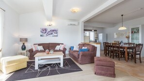 A spacious living area with a sofa set, a coffee table, and a dining table with chairs. There is a kitchen in the background with a refrigerator and a sink.