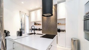 A modern kitchen with white cabinetry, a central island, a black range hood, and a stainless steel trash can. - A Delightful 2-BR/2BA at Saint Michel - Notre Dame (Paris)