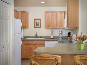 A kitchen with wooden cabinets, a refrigerator, a sink, a blender, a coffee maker, and a dining area with wicker chairs. - Two Bedroom Condo @ Kailua-Kona (Kailua-Kona)