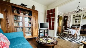 Un salon avec un canapé bleu, un meuble en bois rempli de livres, un téléviseur et un coin repas doté d’un sol à carreaux.