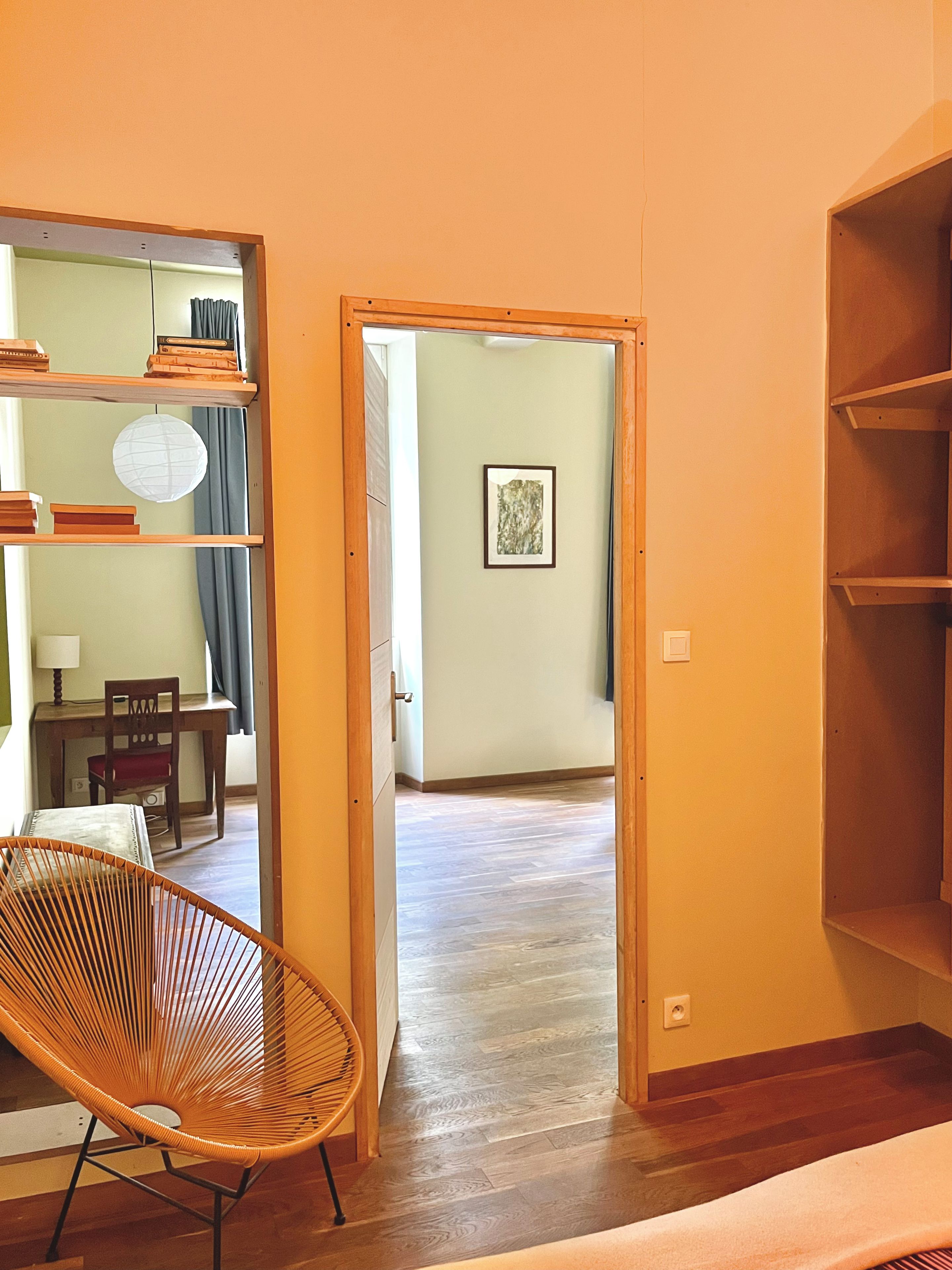 A room with a wooden floor, a chair, a desk with a chair, and a bookshelf.