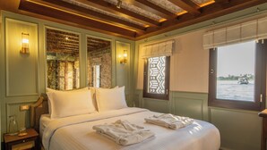 A large bed with white linens, two wall-mounted lamps, and a window with a view of a body of water. - HMC Zahra Dahabyia (Armant)