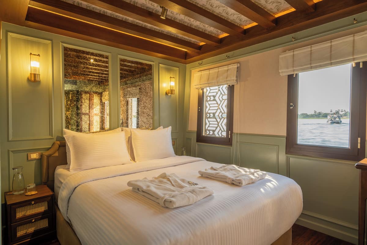 A large bed with white linens, two wall-mounted lamps, and a window with a view of a body of water.