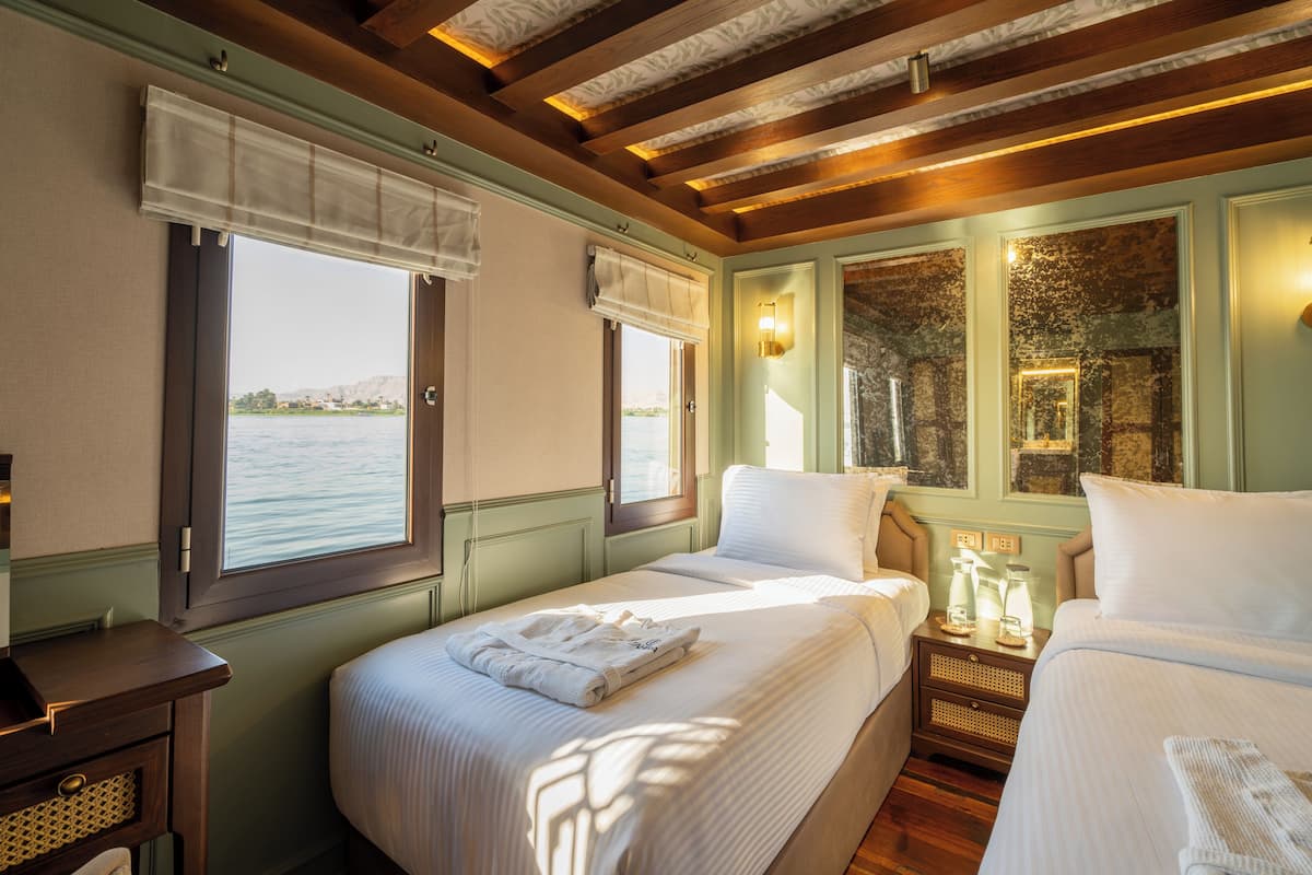 A room with two beds, a wooden ceiling, a view of the water, and a nightstand with a lamp.