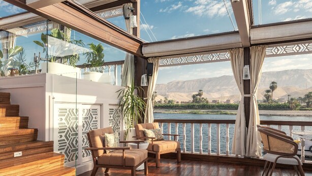 A spacious balcony with wooden flooring, a seating area with cushioned chairs, and a view of a river and mountains. - HMC Zahra Dahabyia (Armant)