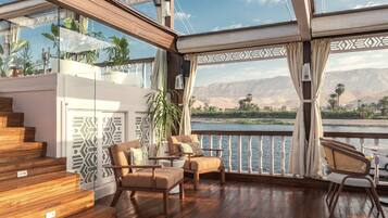 Een ruim balkon met houten vloeren, een zithoek met comfortabele stoelen en uitzicht op een rivier en bergen.