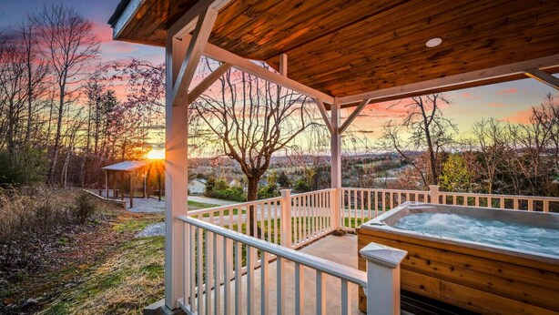 Eine Holzterrasse mit Whirlpool, weißen Geländern und Blick auf einen Sonnenuntergang über einer bewaldeten Landschaft.