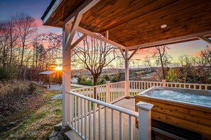 Una terraza de madera con jacuzzi, barandillas blancas y vistas a un bosque al atardecer.