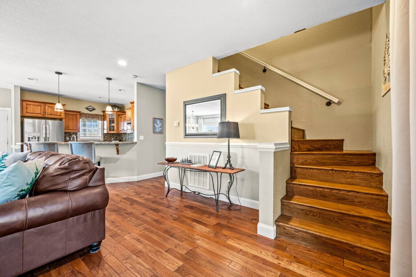 A modern living room with a brown leather couch, wooden flooring, a dining area with a table and chairs, a kitchen in the background, and a wooden staircase.