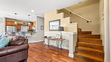 A modern living room with a brown leather couch, wooden flooring, a dining area with a table and chairs, a kitchen in the background, and a wooden staircase.
