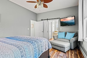 Ein Schlafzimmer mit einem Bett, einem grauen Sessel, einem Deckenventilator und einem Fernseher an der Wand.