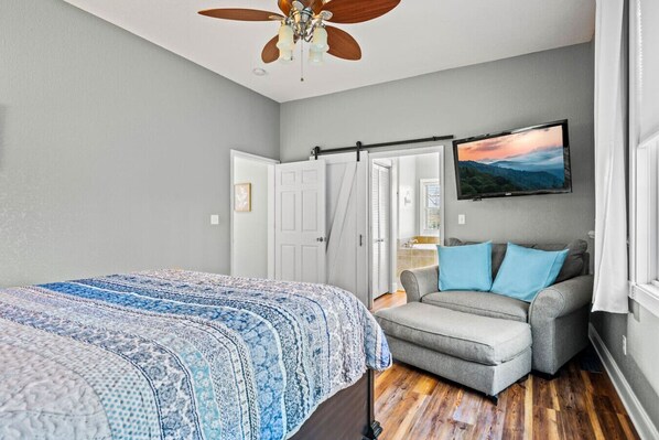 Un dormitorio con cama, un sillón gris, ventilador de techo y televisión en la pared.