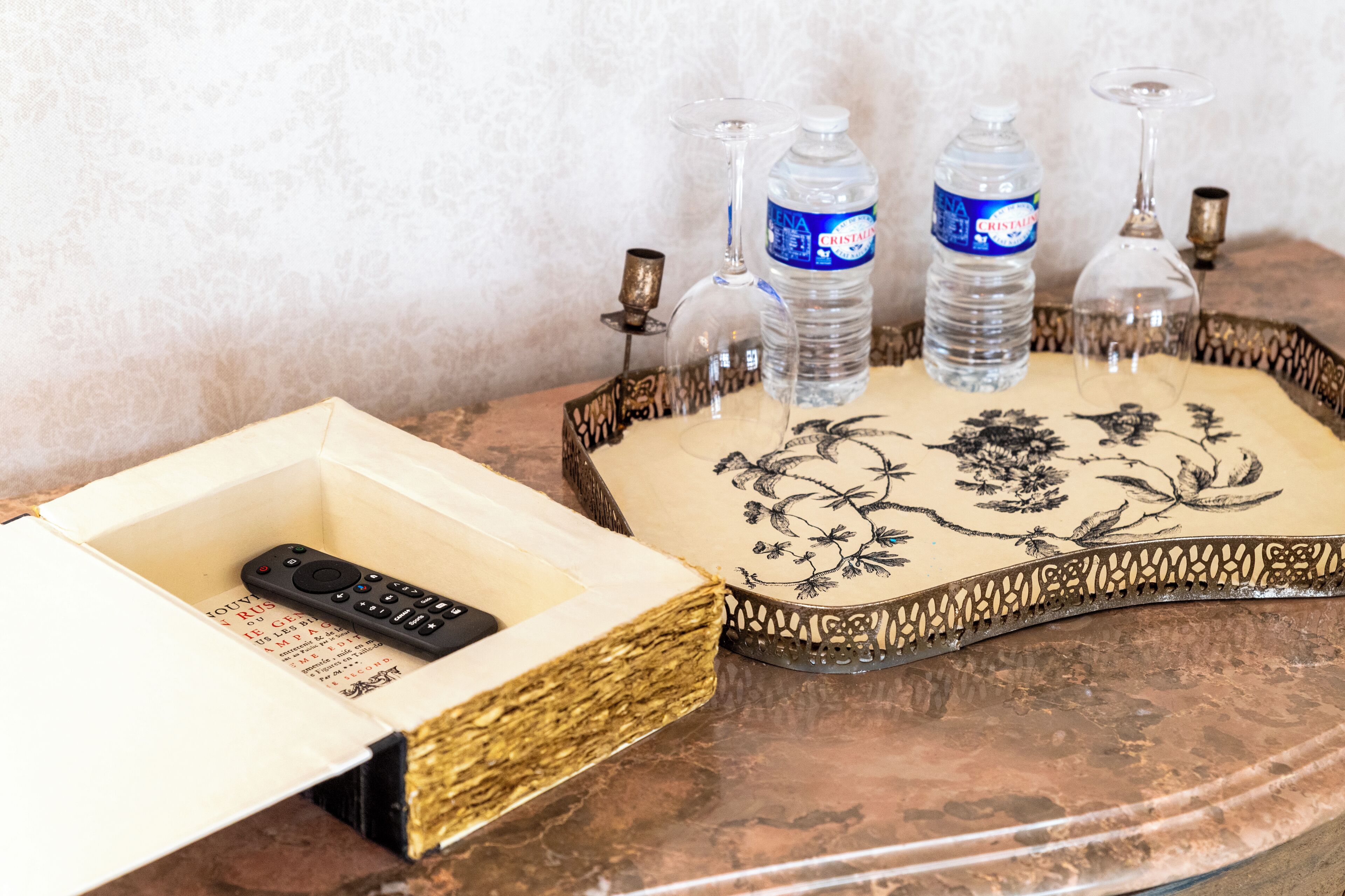 Une télécommande dans une boîte, deux bouteilles d’eau et deux verres à vin sur une table, accompagnés d’un plateau orné de motifs floraux.