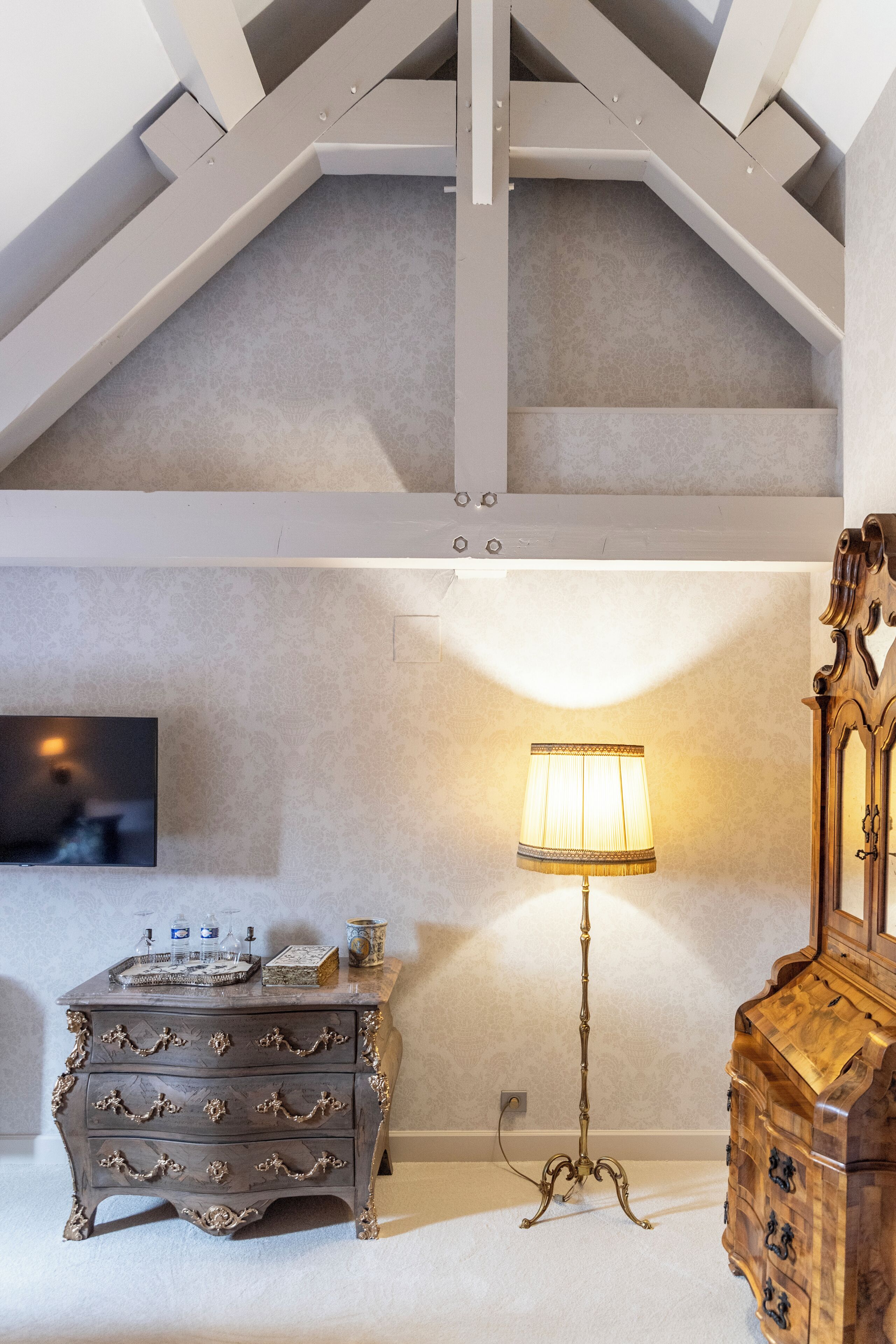 A room with a high ceiling, a wooden dresser, a floor lamp, and a television on the wall.