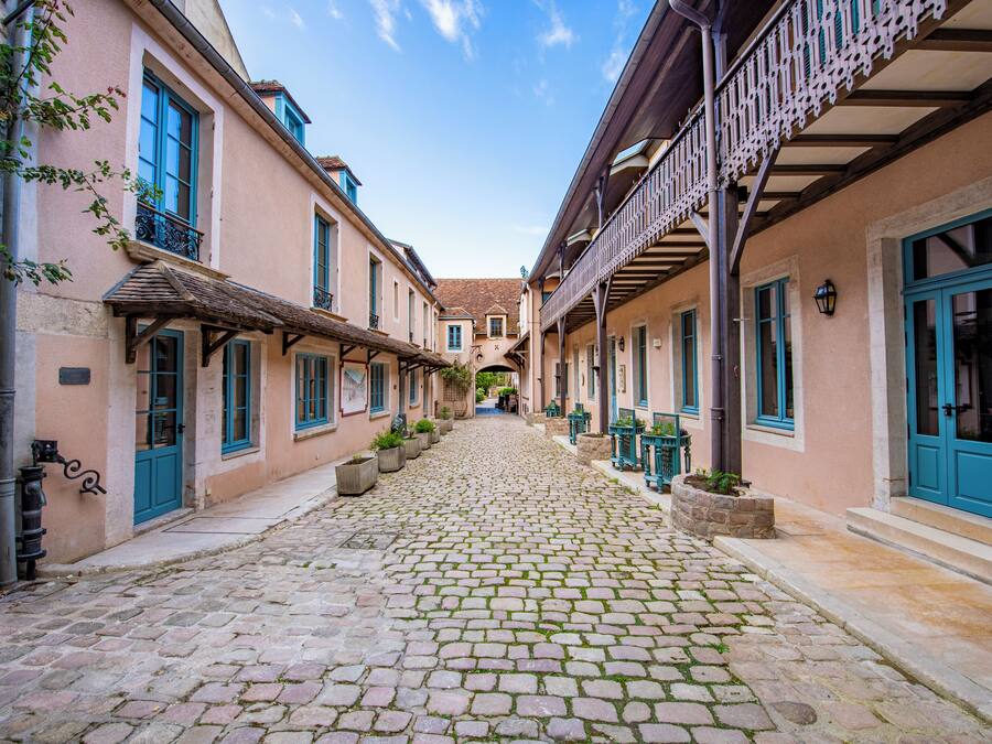 A cobblestone street flanked by buildings with blue doors and windows, a covered walkway, and outdoor seating areas.